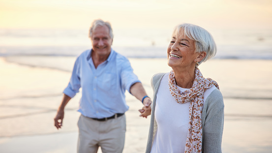 Laughing senior woman leading her husband by the hand while walking together on a sandy beach at sunset in summer