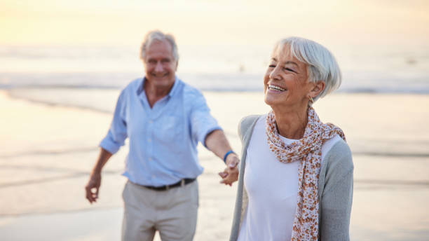 Laughing senior woman leading her husband along a beach at sunset stock photo
