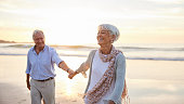 Senior woman laughing while leading her husband along a beach at sunset