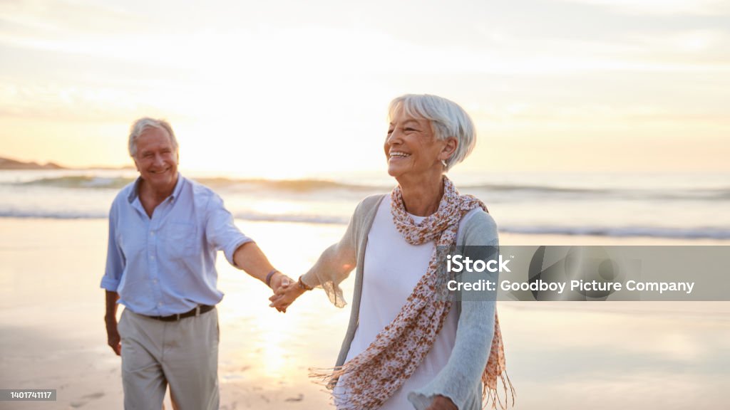 Senior woman laughing while leading her husband along a beach at sunset Laughing senior woman leading her husband by the hand while walking along a sandy beach at sunset in summer Senior Couple Stock Photo