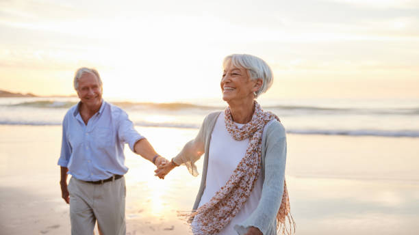 Senior woman laughing while leading her husband along a beach at sunset stock photo