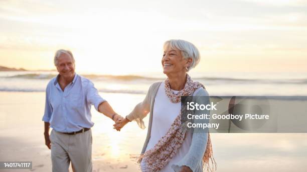 Senior Woman Laughing While Leading Her Husband Along A Beach At Sunset Stock Photo - Download Image Now