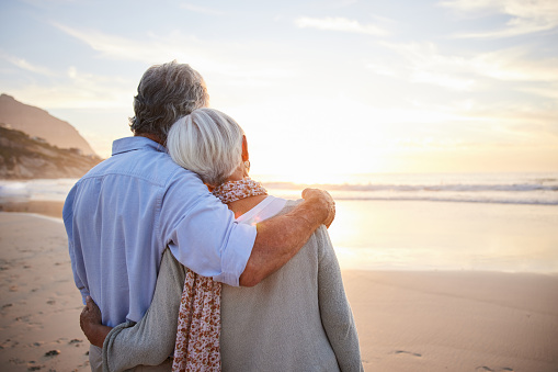 Rear view of a senior couple looking out at the ocean view at sunset while standing arm in arm together on a beach