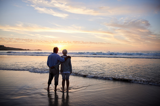 Rear view of a senior couple looking at the sun setting over the ocean while standing hand in hand together on a sandy beach