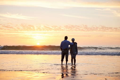 Rear view of a senior couple looking at the sun setting over the ocean while standing arm in arm together on a sandy beach