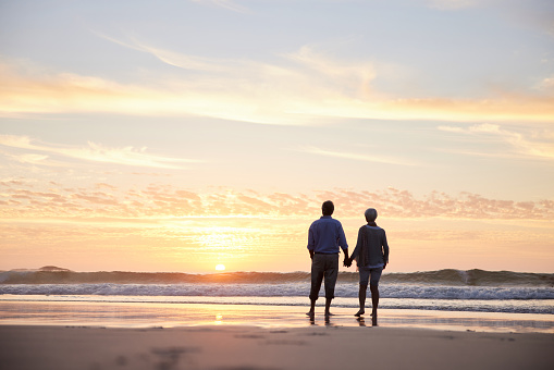 Rear view of a senior couple looking at the sun setting over the ocean while standing holding hands on a sandy beach