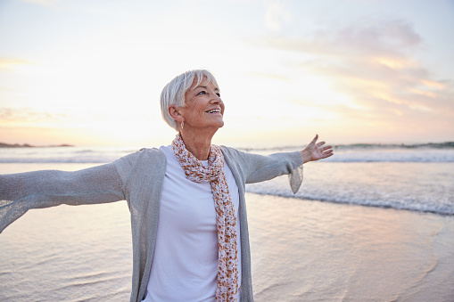Smiling senior woman standing on a sandy beach at sunset with her arms raised to the sky