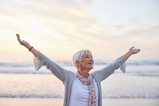 Senior woman smiling while standing with her arms raised to the sky on a sandy beach at sunset in the summertime