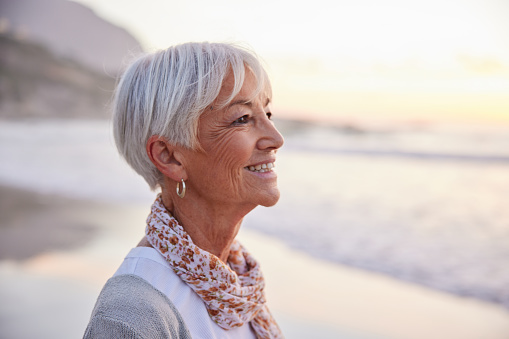 Close-up of a smiling senior woman standing alone on a sandy beach at sunset in summer