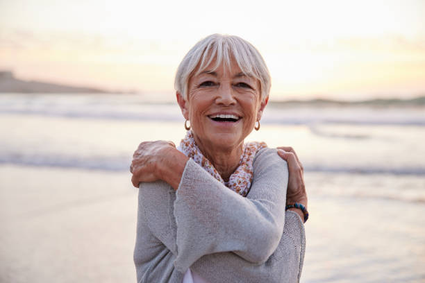 Laughing senior woman hugging herself on a beach at sunset