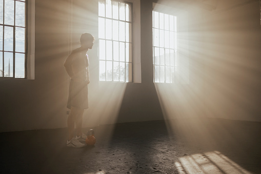 Silhouette of a fit young man standing in the gym with a kettlebell in front of him, getting ready to train while intense rays of light are getting in through the windows