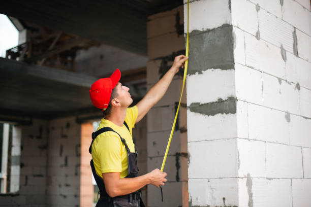 istock Construction worker at construction site measures the length of window opening and brick wall with tape measure. Cottage are made of porous concrete blocks, work clothes - jumpsuit and baseball cap 1401749152