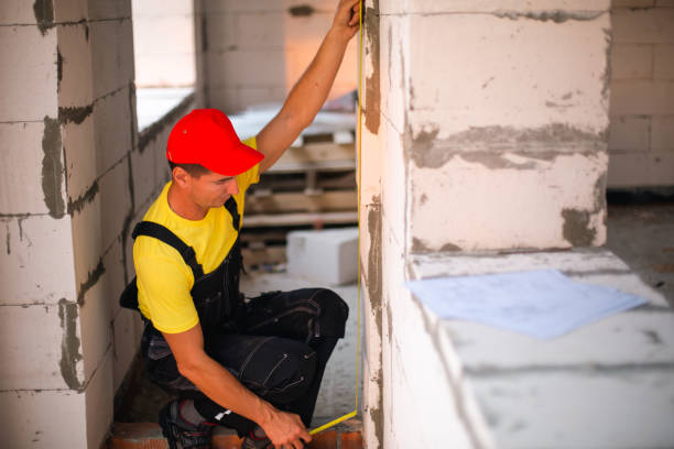 istock Construction worker at construction site measures the length of window opening and brick wall with tape measure. Cottage are made of porous concrete blocks, work clothes - jumpsuit and baseball cap 1401749156