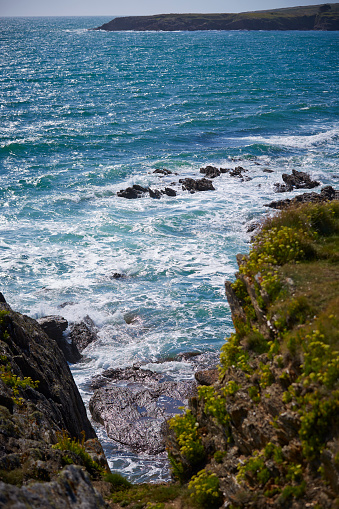 A rough coastline in Brittany, France. The coast is made up of huge rocks and the waves come crashing at them. It is a beautiful day and the water of the Atlantic ocean and sky are both clear blue.