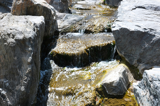 Fresh water creek flow with big boulder rocks and stones close up landscape photo. Rocky river shore natural background image with rough texture. Small waterfall stream with water glimmering in sun.