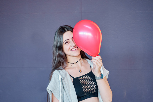 Red air balloon . Pretty young woman with long hair closing half of his face with balloon while smiling and looking at camera