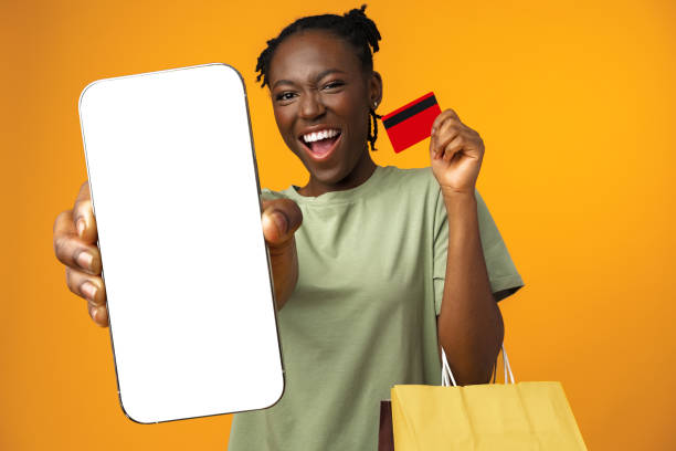 joven afro sonriente sostiene teléfono inteligente y tarjeta de crédito en estudio amarillo - greeting card holding women credit card fotografías e imágenes de stock