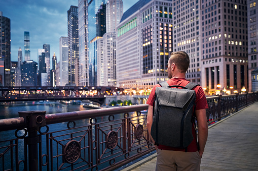 Rear view of man with backpack while walking on bridge and looking around. Illuminated city with skyscrapers at twilight. 