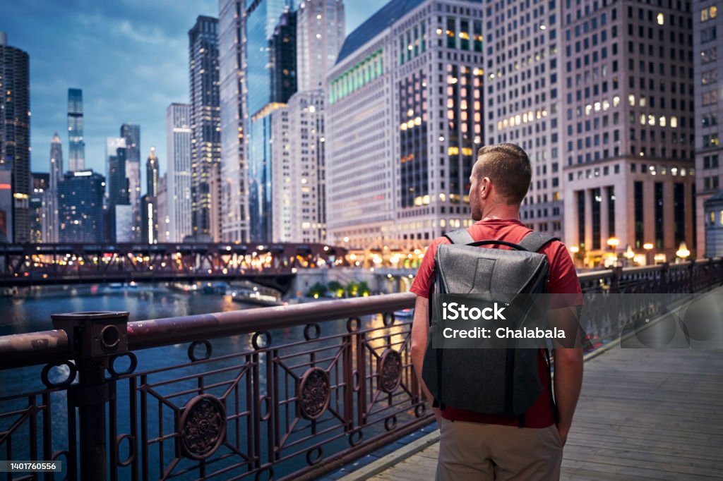 Illuminated city with skyscrapers at twilight Rear view of man with backpack while walking on bridge and looking around. Illuminated city with skyscrapers at twilight. "n Chicago - Illinois Stock Photo