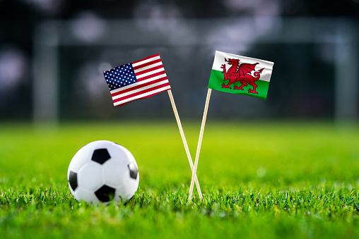 USA vs. Wales, Ahmad Bin Ali, Football match wallpaper, Handmade national flags and soccer ball on green grass. Football stadium in background. Black edit space.