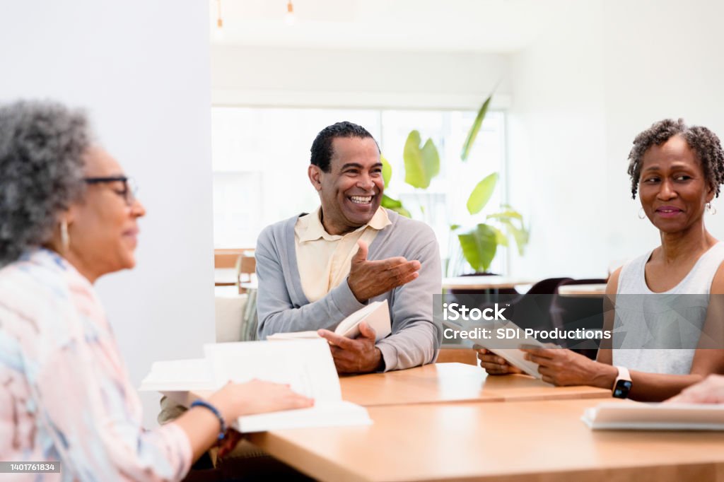 Mature male leader gestures toward unseen person during discussion The mature adult male book club discussion leader smiles and gestures toward an unseen member. Learning Stock Photo