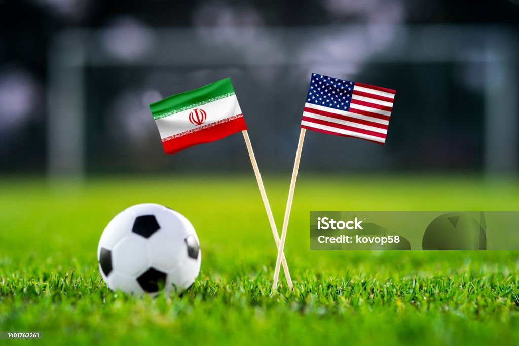 Iran vs. USA, Al Thumama, Football match wallpaper, Handmade national flags and soccer ball on green grass. Football stadium in background. Black edit space. Iran Stock Photo