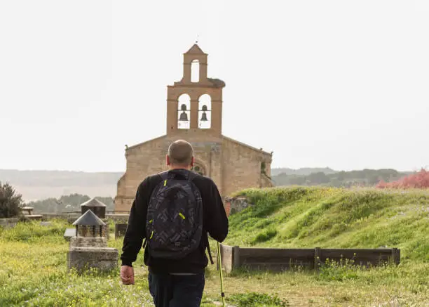In a natural environment on a clear sky day a man walks towards a church on a path, equipped with backpack and trekking pole.