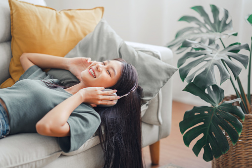 Happy asian woman listening to music from mobile phone while sitting on sofa at homes, Smiling girl relaxing with headphones in morning, Time to relax.