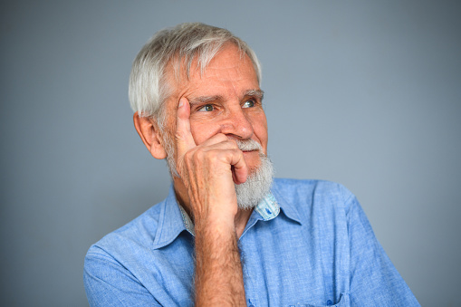 Head shot of thoughtful senior man wearing a blue shirt. He is thinking about the future and holding his head with a hand.