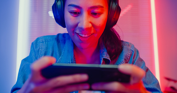 Close up of young Asian woman playing online smartphone video game and broadcast streaming live in neon lights living room at home. Gamer lifestyle concept.