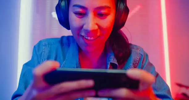 Photo of Close up of young Asian woman playing online smartphone video game and broadcast streaming live in neon lights living room at home.