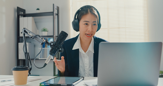 Young Asian businesswoman recording and broadcasting a podcast on her laptop from studio office. Live streaming and broadcasting online concept.