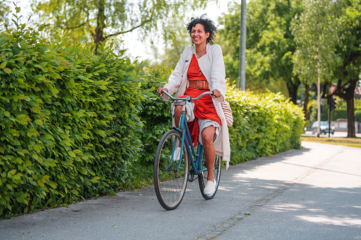 Multiracial woman with curly, black hair, riding a bike to work, looking happy, her skirt pulled up. Full length shot, bright daylight. Green in the background.