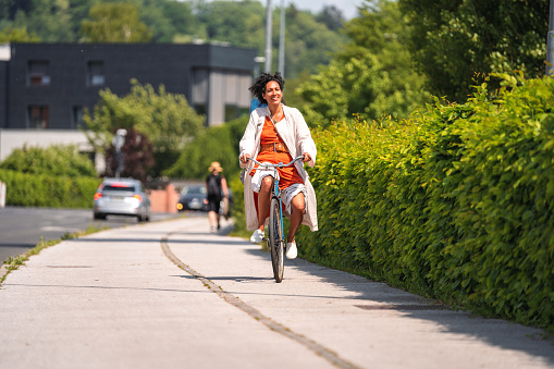Curly, black-haired biracial  woman on a bicycle, riding on the sidewalk next to a busy road with cars in blurred background. Looking relaxed and smiling. Looking away. Full length shot.