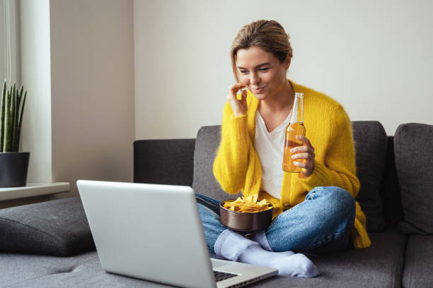 une jeune femme boit de la bière et mange des nachos tout en regardant un film sur son ordinateur portable à la maison - computer chip photos et images de collection