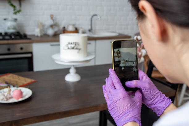 Woman in an apron holds smartphone in her hands and takes pictures of cake. There are chocolate figurines on table. Close-up. Selective focus. Photos about confectioners, food, hobbies. stock photo
