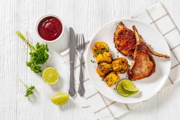 roasted tomahawk pork chops, bone-in steak with grilled corn disks and lime on white plate on white wooden table with barbeque sauce, horizontal view from above, flat lay