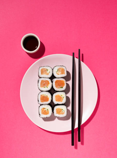 japanese sushi roll with salmon, cucumber and soy sauce with chopsticks in the plate on pink background with hard shadow. concept food photography. top view and copy space - sushischotel stockfoto's en -beelden