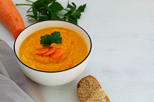 white bowl of tasty orange carrot cream soup with bread and herbs on white background.Spring vegetable Homemade Carrot cream-soup with parsley on table.copy space.