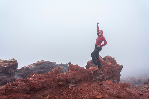 Mature women standing on the red rocks of recent eruption of volcano Eldfell, on island of Heimaey in Vestmannaeyjar islands with raised one arm.