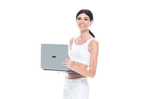 Front view of aged 20-29 years old who is beautiful caucasian female athlete standing in front of white background wearing leggings who is smiling who is working and using laptop