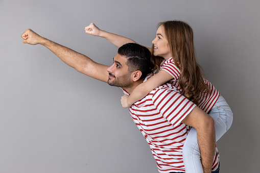 Cheerful daddy piggybacking daughter, making flying superhero hand, fist forward gesture, little girl riding fathers back, playing active game together. Indoor studio shot isolated on gray background.