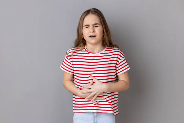 Little girl clutching belly, feeling discomfort in stomach, sick child suffering constipation cramps Portrait of little girl wearing striped T-shirt clutching belly, feeling discomfort or pain in stomach, sick child suffering constipation cramps. Indoor studio shot isolated on gray background. nausea stock pictures, royalty-free photos & images