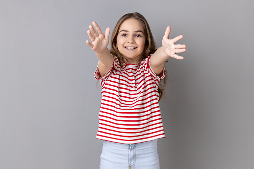 Come into my arms. Portrait of smiling happy little girl wearing striped T-shirt outstretching hands to embrace, giving free hugs and welcoming. Indoor studio shot isolated on gray background.