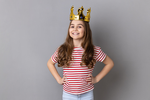 Portrait of satisfied arrogance little girl princess wearing striped T-shirt and golden diadem crown, looking at camera keeps hands on hips. Indoor studio shot isolated on gray background.