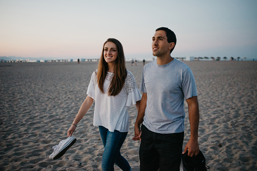 A Hispanic man with his smiling girlfriend are strolling on a sandy beach near the Balearic sea in Spain in the evening. A couple of tourists on a date at twilight in Valencia.