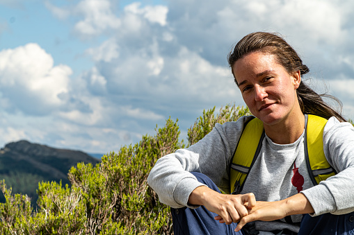 Blonde woman with wavy hair, sitting on a rock while resting on a mountain hiking trail.