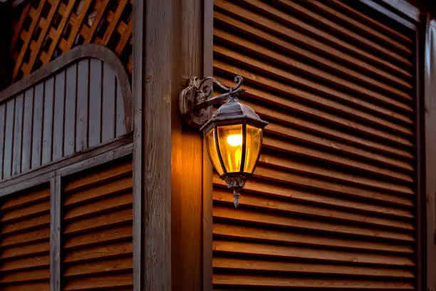 iron forged retro lantern with a glass shade and an electric light bulb on a wooden wall made of planks, wooden shutters architecture with evening lighting with a warm glow, closeup.