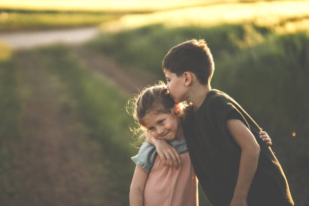 Two kids, brother hugging and kissing his little sister on the meadow outdoors in sunset.
