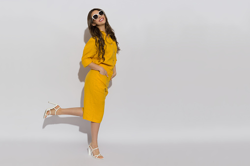 Happy young woman in sunglasses, linen shirt, shorts and high heels poses on a one leg. Full length studio shot against white background